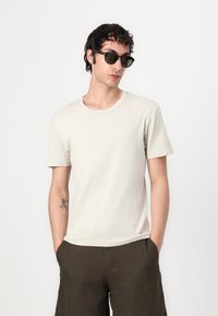 Beige crew neck t-shirt in a soft fabric, paired with dark brown shorts. Model wearing black sunglasses, hands in pockets, stands against a light background.
