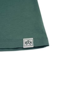 Tummanvihreä puuvillapaita, jossa on sileä pinta. Paidan helman kohdalla on valkoinen etiketti, jossa on musta logo ommeltuna.