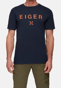 Navy blue cotton t-paita, jossa on oranssi "EIGER" -teksti painettuna rinnassa. Klassinen pyöreä kaula-aukko, lyhyet hihat ja normaali istuvuus.