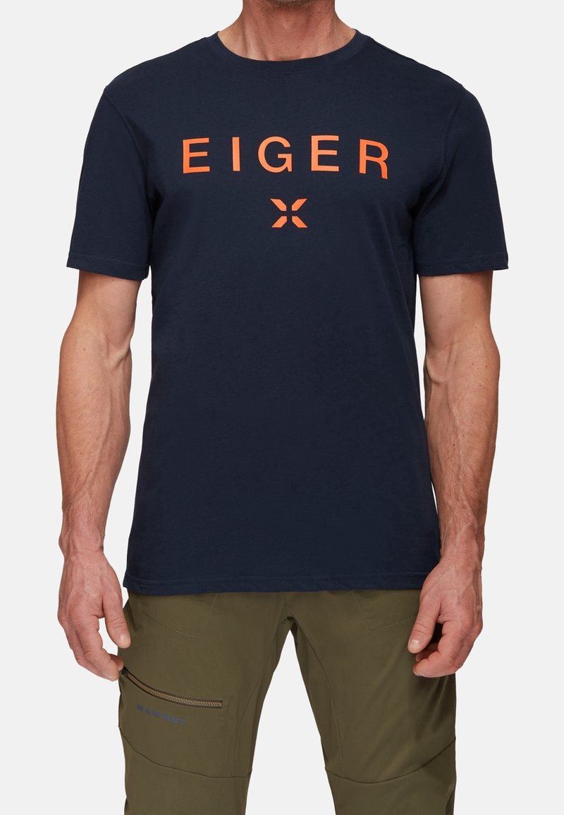 Navy blue cotton t-paita, jossa on oranssi "EIGER" -teksti painettuna rinnassa. Klassinen pyöreä kaula-aukko, lyhyet hihat ja normaali istuvuus.
