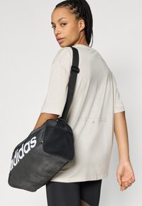 Μαύρη τσάντα duffel με λευκό λογότυπο "adidas", με υφή και λεπτομέρειες από δίχτυ. Φορεμένη με φαρδιά μπεζ μπλούζα και μαύρα κολάν.