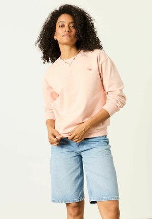 Frau mit lockigem Haar, die einen hellrosa Pullover, eine Perlenkette und hellblaue Jeans-Shorts knielang trägt, steht vor einem einfarbigen Hintergrund.