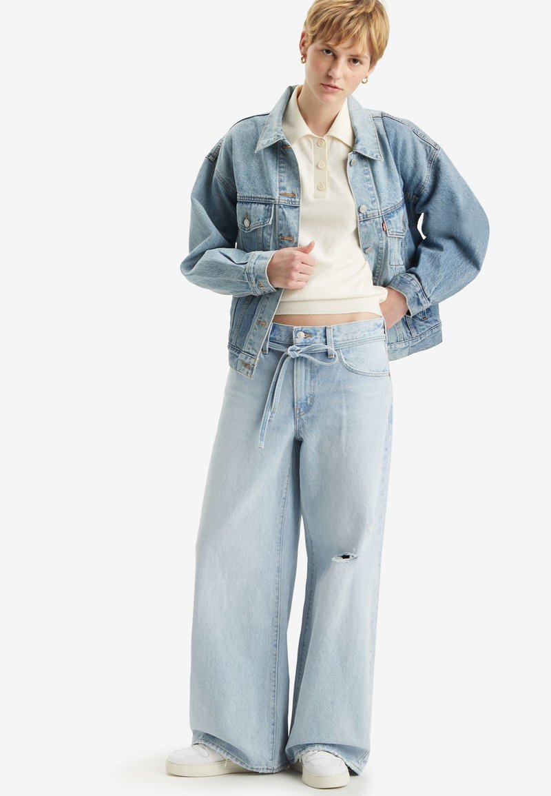 Veste en denim bleu clair et jean large avec cordon de serrage, associés à un polo crème et des baskets blanches. Détails effilochés sur le jean.