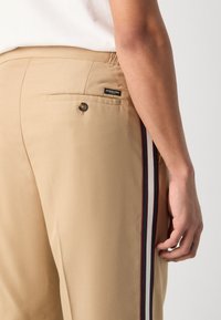 Beige skreddersydde bukser med elastisk midje, sidestroppdetaljer i marineblått, rødt og hvitt, samt en baklomme med knappelukking.