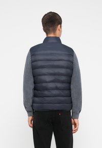 Mann in einer marineblauen gepolsterten Weste über grauen Strickärmel, von hinten betrachtet, mit schwarzen Jeans vor einem schlichten weißen Hintergrund.