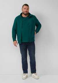 Man met een baard en vastgebonden haar, gekleed in een turquoise ritsjack, een bijpassend T-shirt, donkere jeans en witte sneakers, staande tegen een grijze muur.