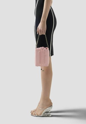 Femme en robe noire avec des finitions blanches tenant un sac à main rose à cordon, portant des sandales à talons transparents aux talons sculptés.