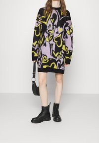 Abito in maglia con motivo a cuori multicolore in nero, viola e giallo; maniche a costine; abbinato a stivali combat neri con lacci e una piccola borsa.