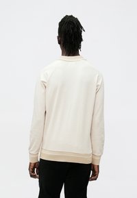 Šviesiai smėlio spalvos džemperis su tekstūruota medžiaga, ribotomis rankogaliais ir apvalia iškirpte. Nugaros vaizdas atskleidžia laisvą siluetą ir paprastus dizaino elementus.