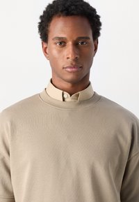 Beige svetr s vysokým límcem, pod kterým je viditelná košile s knoflíky. Hladká textura a uvolněný střih.