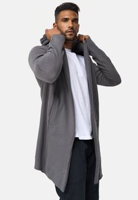 Sudadera larga gris con abertura frontal, gran capucha y dos bolsillos laterales, llevada sobre una camiseta blanca y pantalones negros. Tejido suave y texturizado.
