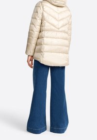 Světle béžová péřová bunda s kapucí, s chevronovým prošíváním a mírně delším zadním dílem. V kombinaci s širokými modrými džínami.