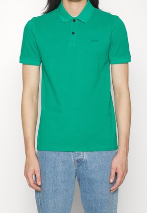 Person trägt ein grünes kurzärmeliges Poloshirt mit schwarzen Knöpfen und hellblauen Jeans, steht vor einem schlichten weißen Hintergrund.