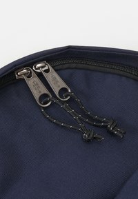 Närbild av en marinblå tygväska med dubbla silverdragkedjor och svarta snoddar. Texturerad yta med sömdetaljer längs kanterna.