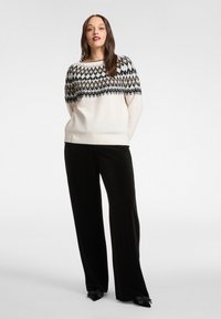 Sweter z dzianiny w kremowym kolorze z geometrycznym czarno-beżowym wzorem na karczku. Charakteryzuje się luźnym krojem i prążkowanym wykończeniem dołu. Połączony z szerokimi czarnymi spodniami.