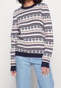 Pull en tricot présentant un motif bleu marine avec des rayures roses et blanches, des flocons de neige et des motifs géométriques. Poignets et ourlet côtelés.