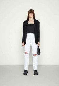 Blazer oversized preto, top preto sem alças, jeans brancos rasgados, botas pretas chunky. A segurar uma pequena bolsa preta. Fundo simples.