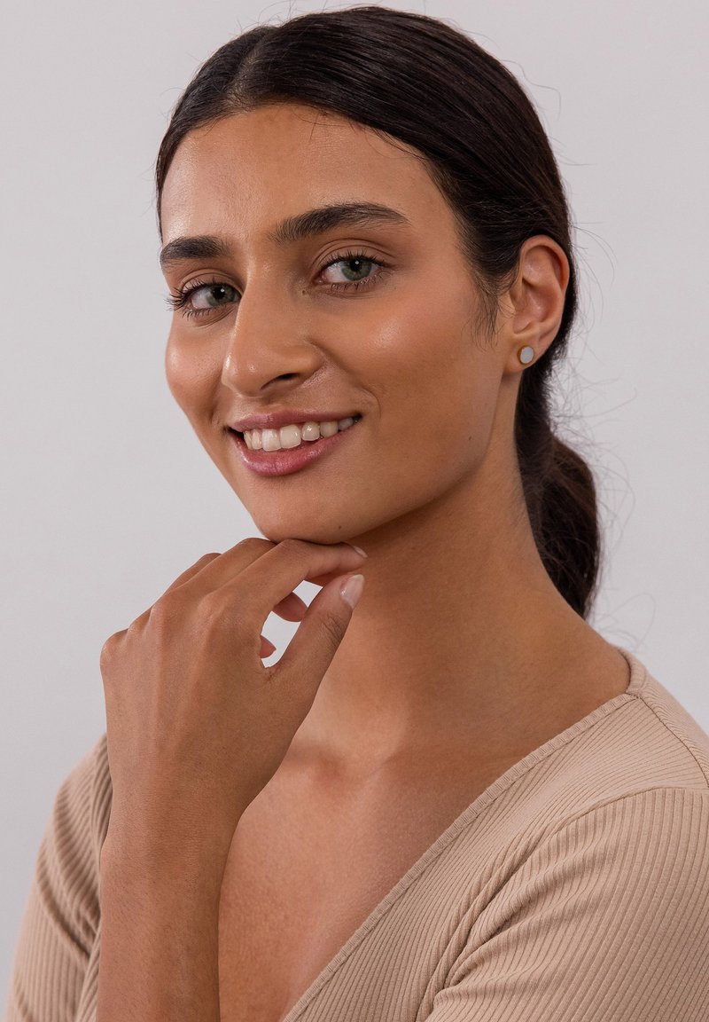 Hellbraun gerippter Oberteil, frontal blickend, Hand am Kinn. Klare Haut und dezentes Make-up, minimale runde Ohrringe, sanfte Beleuchtung, neutraler Hintergrund.