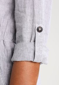 Manche retroussée sur une chemise rayée grise et blanche sécurisée par une patte boutonnée noire au-dessus de l'avant-bras.