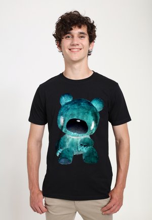 Smilende ung mand med krøllet hår iført en sort T-shirt med et stort blågrønt bamsemotiv på forsiden.