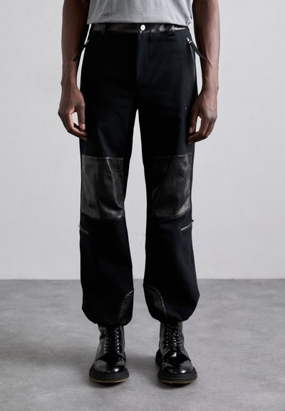 Pantalons noirs avec des empiècements en cuir contrastants, des poches zippées et une coupe droite. Portés avec des bottes noires à lacets.
