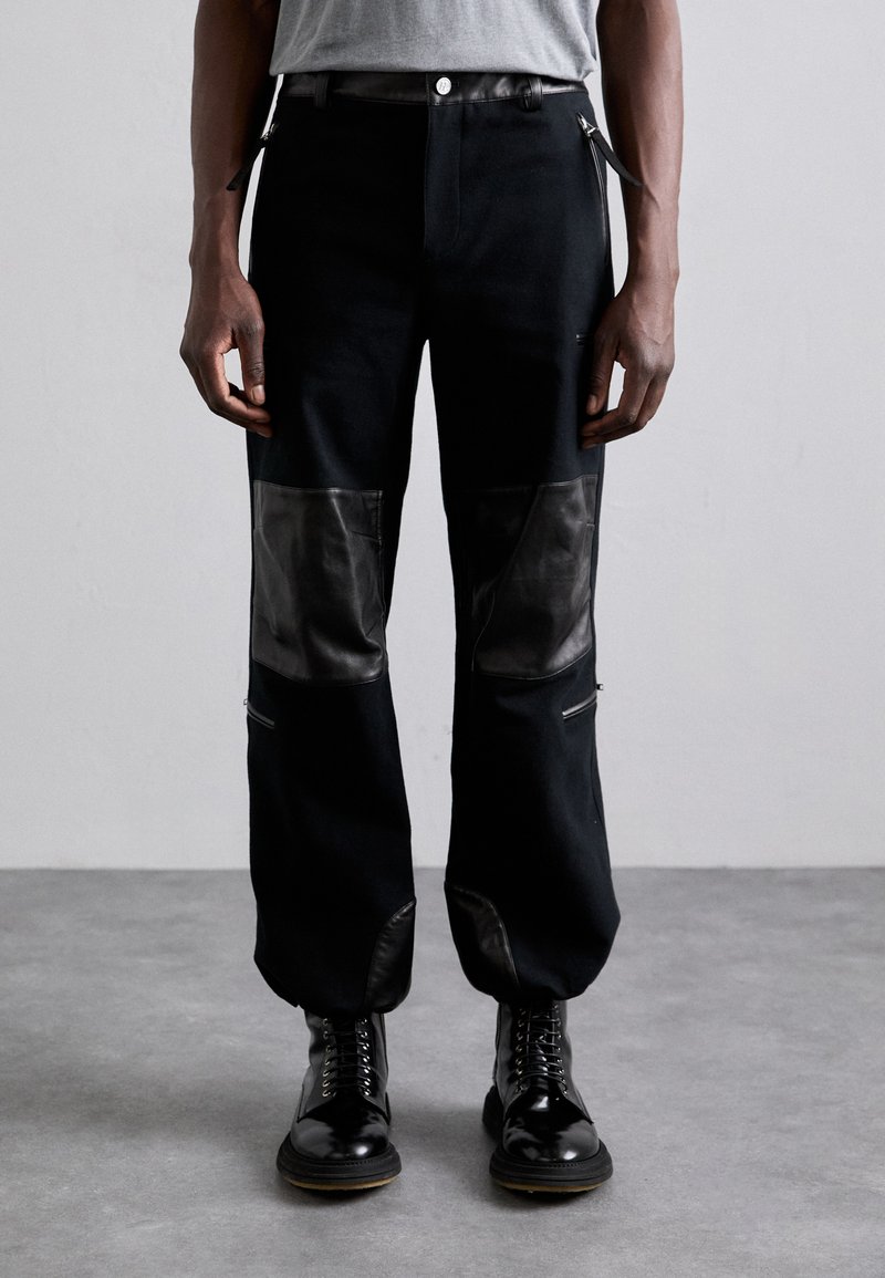 Schwarze Hose mit kontrastierenden Lederpatches, Reißverschlusstaschen und geradem Schnitt. Getragen mit schwarzen Schnürstiefeln.