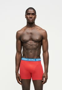 Homme musclé debout, portant un boxer rouge avec une ceinture bleue avec le texte "BJÖRN BORG", fond neutre.