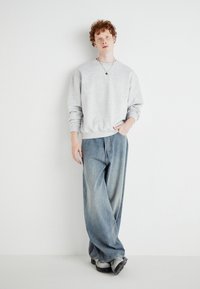 Sweatshirt gris à coupe décontractée, associé à un pantalon en jean large bleu clair. Accessoirisé avec un collier pendentif, se tenant devant un fond uni.