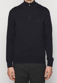 Stickad mörkblå pullover med hög krage och kvartzipper, med en diskret logotyp på bröstet. Mjuk textur.