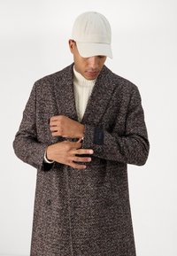 Abrigo marrón de tweed con tejido texturizado, frontal de doble botonadura, botones negros y etiqueta visible en el puño. Suéter debajo.