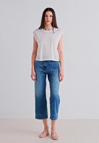 Cropa blanca de manga corta; vaqueros anchos de mezclilla azul con dobladillos deshilachados; zapatos bajos beige claro; fondo liso.