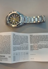 Orologio in acciaio inossidabile argentato con quadrante nero, dettagli gialli e tre sottoquadranti. Accompagnato da una scheda di garanzia piegata con testo multilingue.