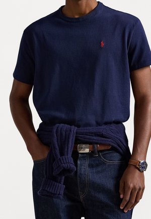 Homme portant une chemise bleu marine à manches courtes avec un logo rouge, un jean foncé, une montre en cuir marron et un pull bleu marine noué autour de la taille.