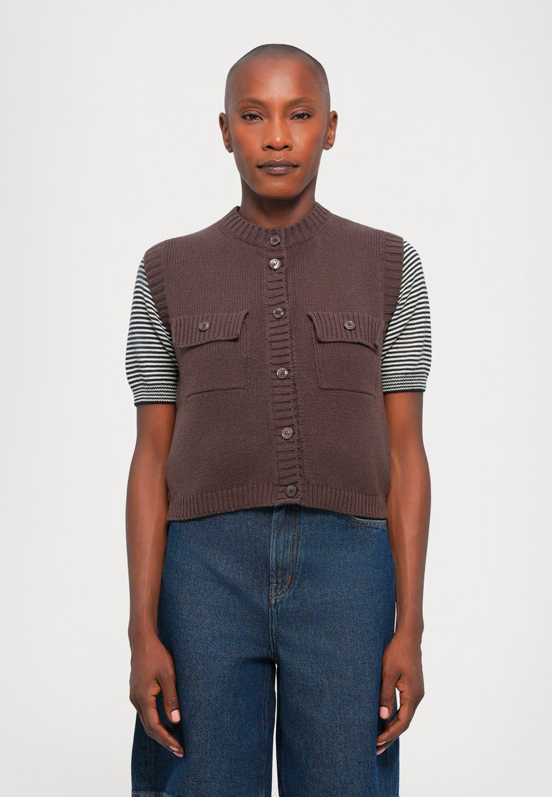 Gilet lavorato a maglia marrone con scollo rotondo, chiusura a bottoni e due tasche sul petto. Le maniche corte a righe aggiungono un contrasto. Indossato sopra jeans a vita alta.