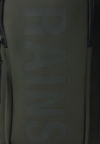 Mörkgrön vattentät ryggsäck med ett stort präglat "RAINS"-logotyp. Slät yta och kontrasterande dragkedjedetaljer är påtagliga egenskaper.