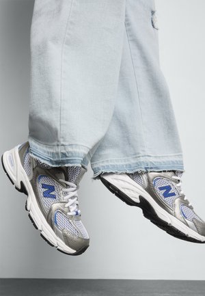 Baskets sportives avec une tige en mesh respirant gris et blanc, rehaussées de accents bleus, posées sous un pantalon en denim bleu clair avec des bords effilochés.