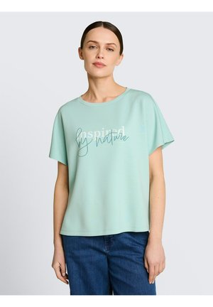 Frau trägt ein hellblaues T-Shirt mit dem Aufdruck "Inspired by nature" und blaue Jeans, steht vor einem einfarbigen hellgrauen Hintergrund.