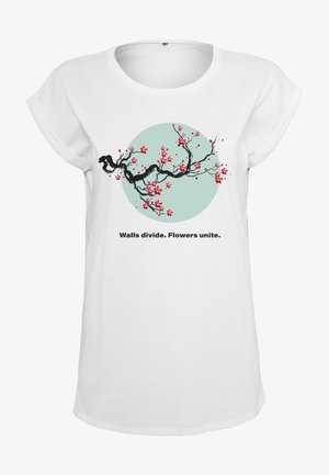 Fehér, rövid ujjú póló, amelyen egy faág piros virágokkal a mentazöld kör háttér előtt látható. Szöveg: "A falak elválasztanak. A virágok egyesítenek."