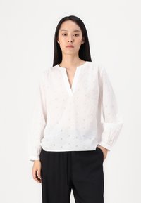 Blusa blanca de manga larga con escote en V, que presenta un patrón floral de ojales y puños fruncidos. Combinada con pantalones negros.
