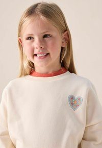 Sweatshirt couleur crème avec un col en corail côtelé, présente un design en forme de cœur avec des motifs floraux dans des teintes pastel. Texture douce.