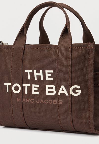 Hnedá plátenná taška s krátkymi rúčkami a bielym nápisom "THE TOTE BAG MARC JACOBS" natlačeným na prednej strane.
