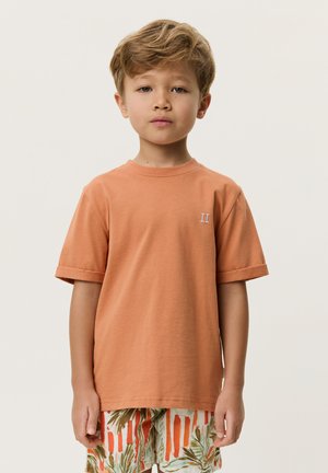 Jong kind met kort lichtbruin haar, draagt een oranje shirt met korte mouwen en een korte broek met patroon, staand tegen een effen lichte achtergrond.
