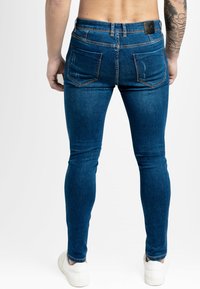Jeans de mezclilla ajustados de color azul con una textura suave, bolsillos traseros, ligero desgastado y una etiqueta negra en la cinturilla. Zapatillas blancas visibles.