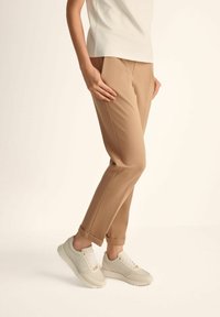 Beżowe spódnice wąsko zwężane o gładkiej fakturze i delikatnych plisach, zestawione z jasnymi trampkami z białymi sznurówkami i jasną podeszwą.