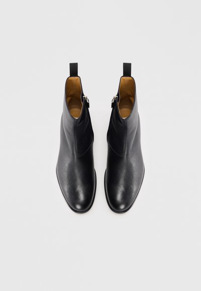 Botas de tobillo de cuero negro con un acabado texturizado, punta afilada y cierre de cremallera lateral. Forro interior suave con tiradores en la parte trasera.