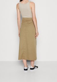 Vrouw gekleed in een beige tanktop, een hooggelegen tan midi rokje met zijzakken, en zwart-witte Adidas sneakers, staande tegen een effen achtergrond.