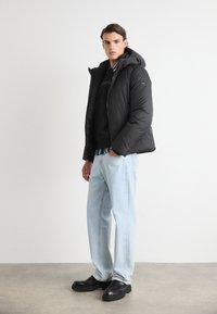 Svart dunjakke med hette, med glidelås, brukt over en svart genser, sammen med lyseblå jeans og svarte sko.