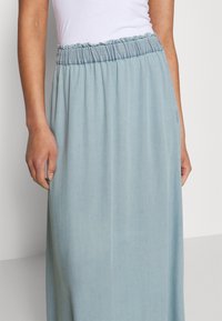 Kvinna klädd i en ljusblå, elastisk midijurken och en vit ärmlös topp, stående med händerna avslappnat vid sidorna.