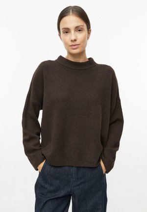 Vrouw met donkerbruine haren in een lage knot, draagt een donkerbruine trui en donkerblauwe spijkerbroek, handen in de zakken, kijkt naar voren.