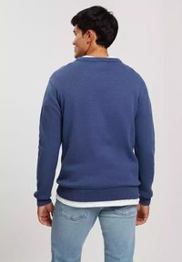 Nieprzemakalny, granatowy sweter z ribbowym kołnierzem i mankietami, o luźnym kroju. Noszony na białej koszuli, z widocznymi detalami szwów na plecach.